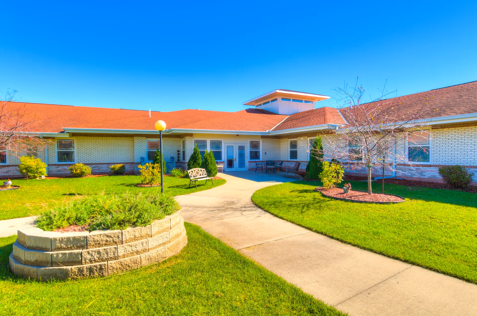 Sunlit courtyard and entrance of a single-story senior living facility with a paved walkway, benches, and landscaped lawn.