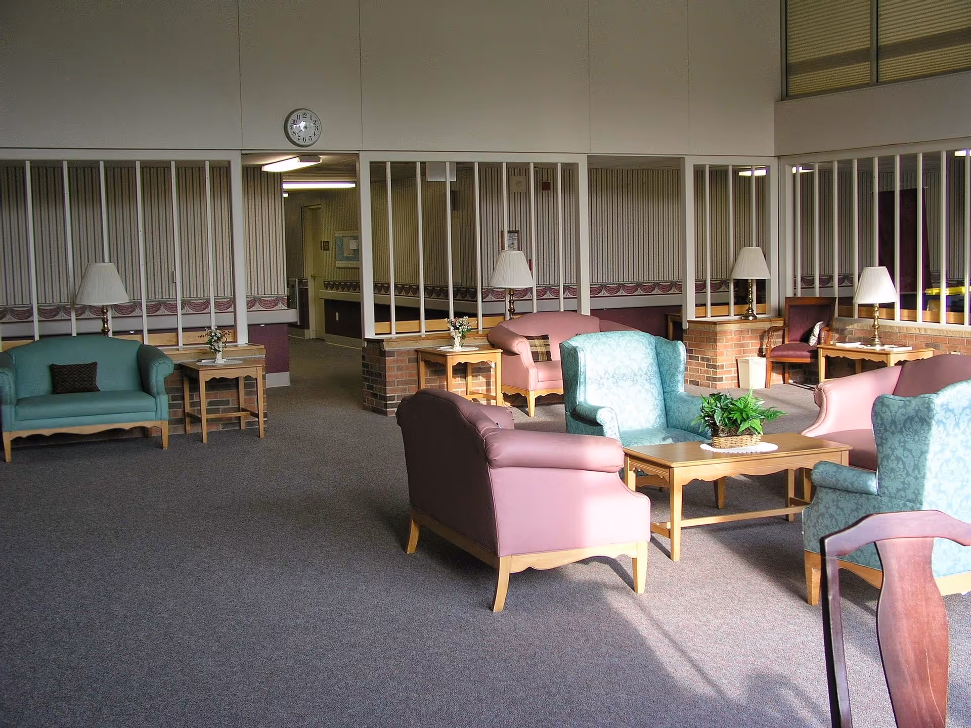 A spacious senior living facility common area with multiple upholstered chairs and sofas arranged around wooden coffee tables. The room has carpeted floors, table lamps, and decorative plants. There are large windows with vertical blinds and a clock on the wall above a hallway entrance.