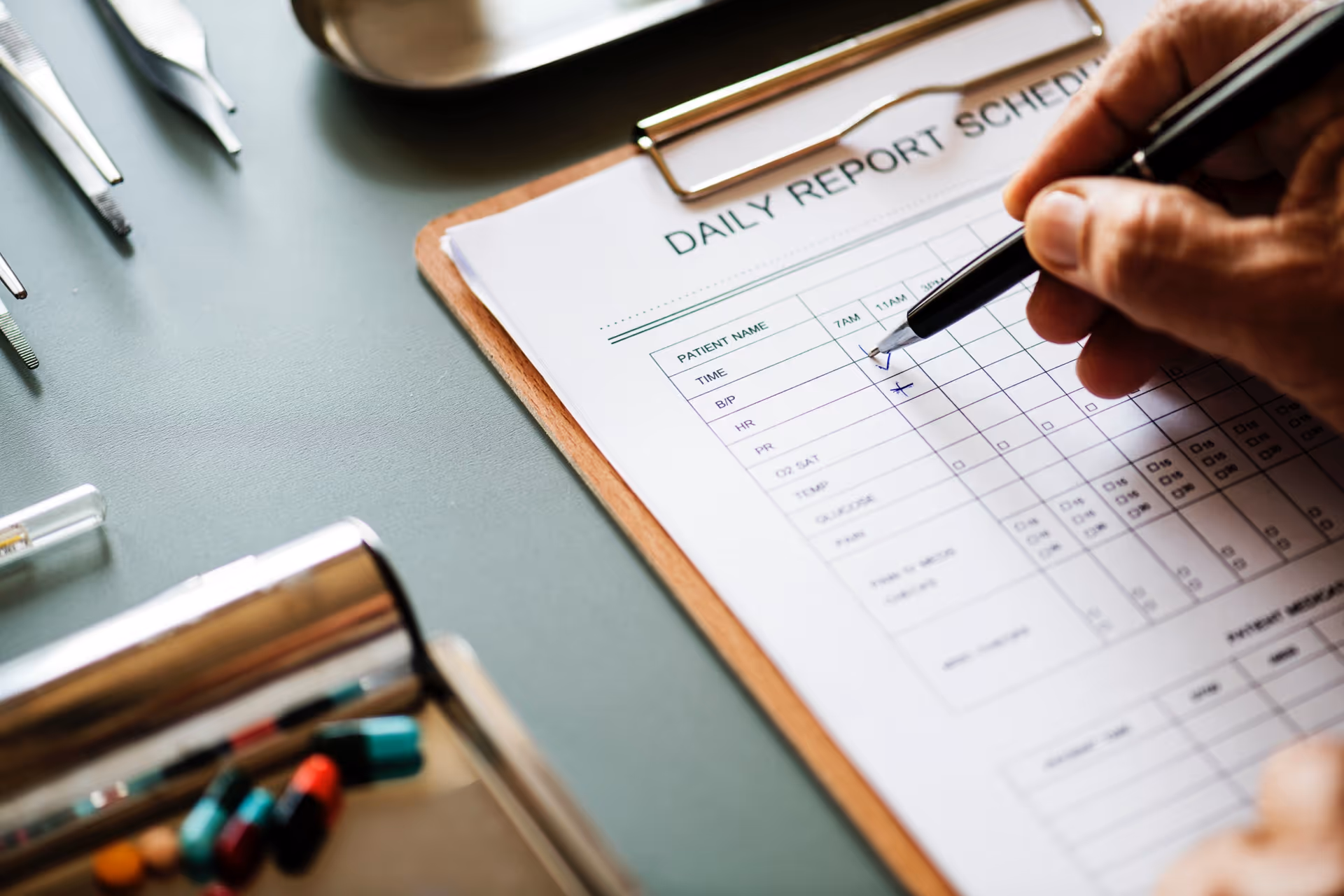 Close up of a hand writing on a daily report schedule form beside medical supplies.