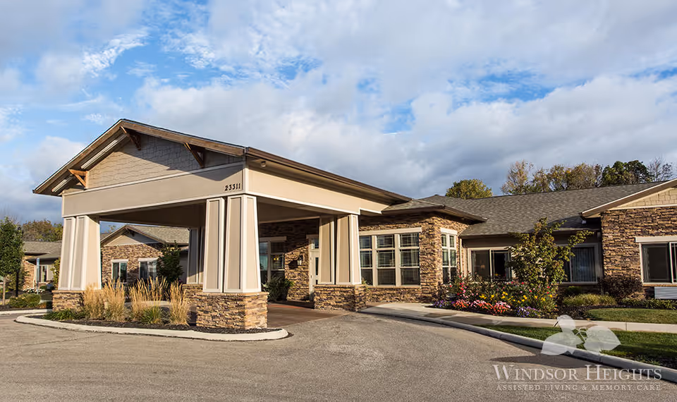 Covered front entrance and porte-cochère of a single-story assisted living building with stone facade and landscaped beds under a partly cloudy sky.