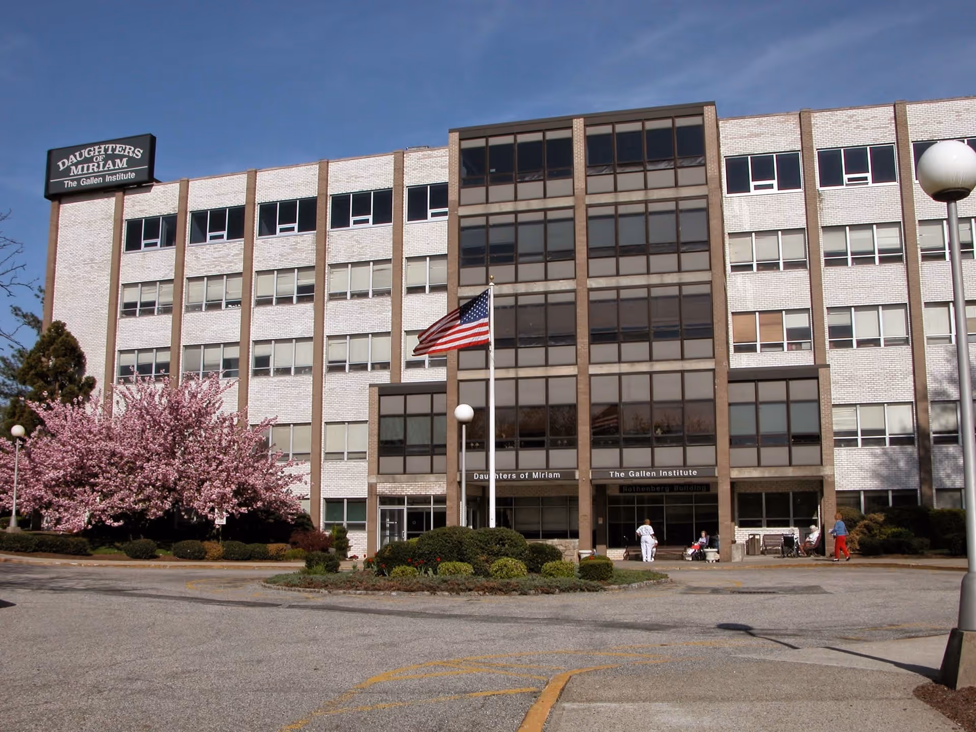 Exterior view of a multi-story brick building with large windows, an American flag on a flagpole in front, and blooming pink trees on the left side. Several people are near the entrance, and the building has signs reading 'Daughters of Miriam' and 'The Gallen Institute'.