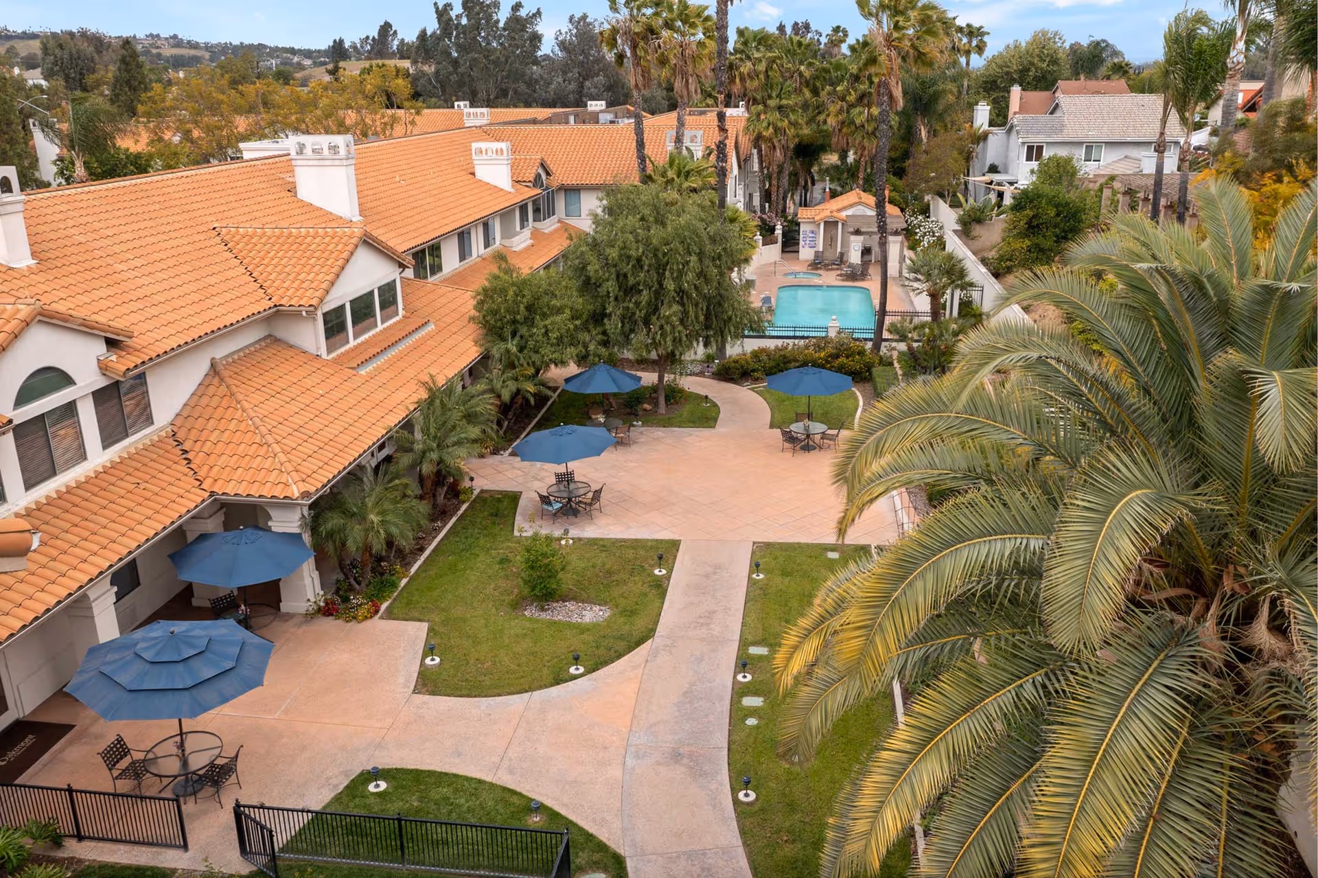 Aerial view of an outdoor courtyard area at Oakmont of Chino Hills featuring a tiled walkway, green lawns, several blue patio umbrellas with tables and chairs, surrounded by buildings with terracotta tiled roofs and palm trees. A swimming pool is visible in the background.