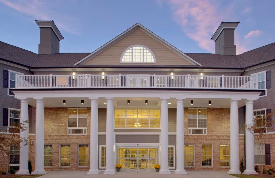 Front exterior view of a senior living facility building with large white columns, multiple windows, and a balcony railing under a twilight sky.