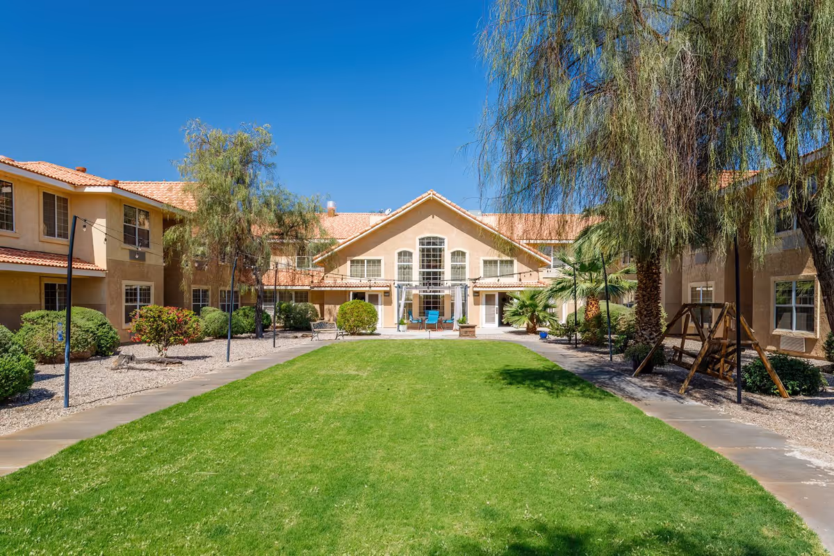 Outdoor courtyard area of Prestige Assisted Living At Lake Havasu featuring a well-maintained green lawn, surrounded by a two-story building with beige walls and red tile roofing. The courtyard includes trees, bushes, a wooden swing, benches, and a seating area with blue chairs under a pergola.