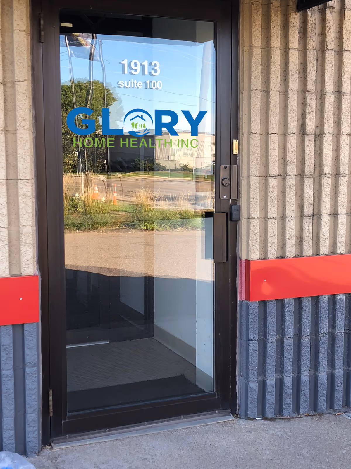 Glass door entrance displaying Glory Home Health Inc logo on a brick building.