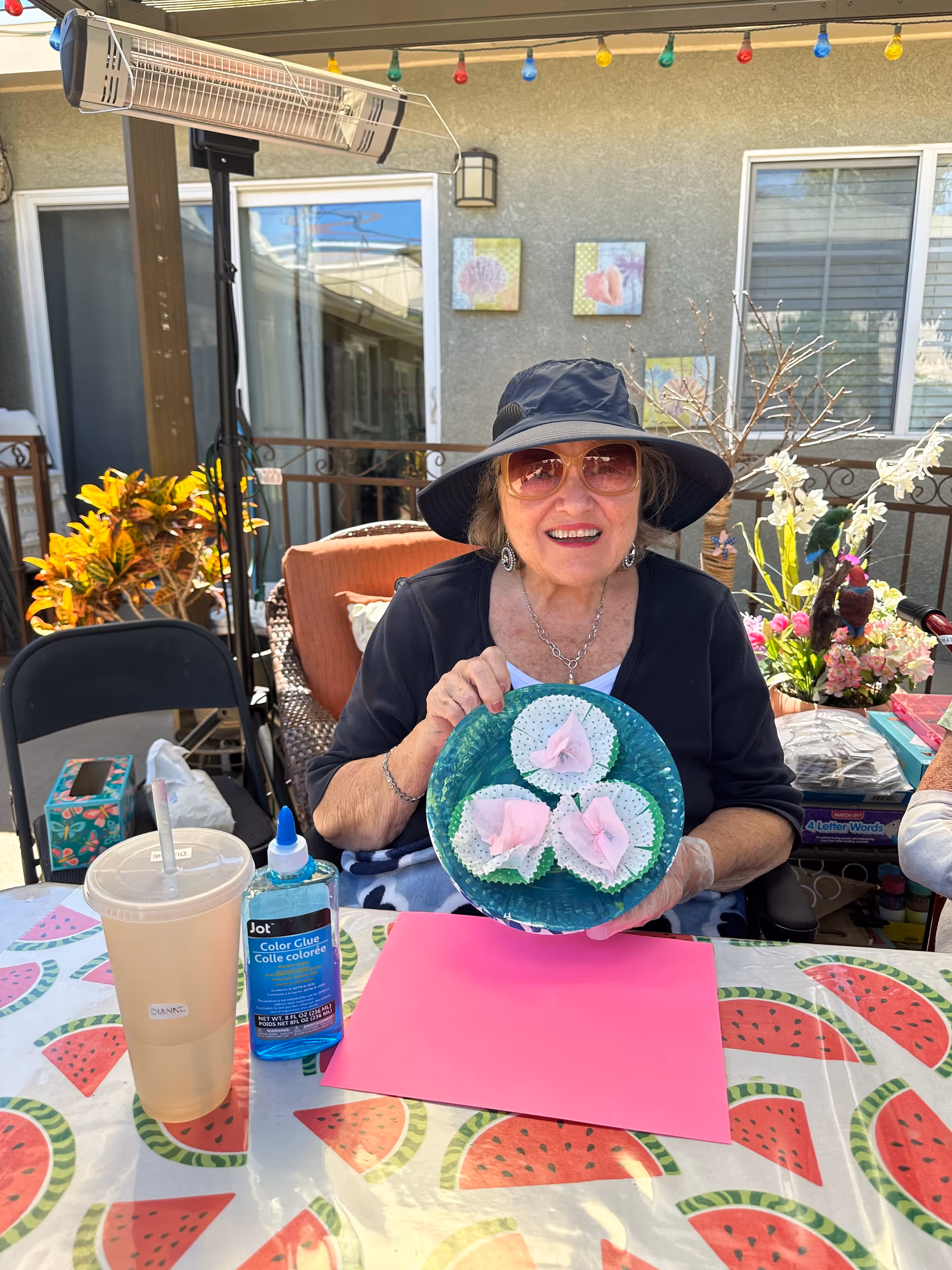 An elderly woman wearing a black wide-brimmed hat and sunglasses is sitting at a table outdoors. She is smiling and holding a blue plate with three pink flower crafts made from paper and cupcake liners. The table has a watermelon-patterned tablecloth and contains a large drink with a straw, a bottle of blue color glue, and a pink sheet of paper. Behind her are plants, flowers, and a building wall with two small framed pictures and a window.