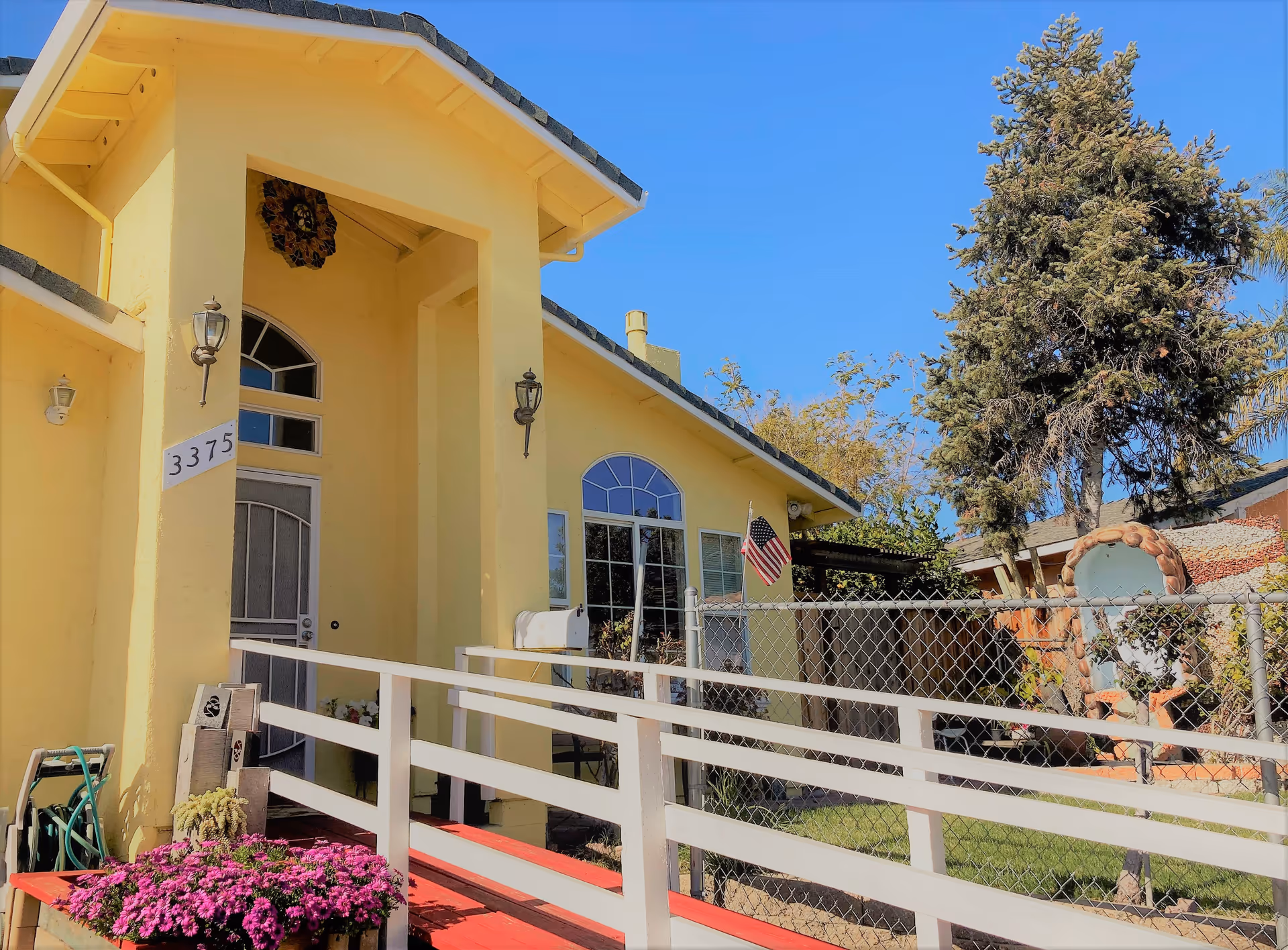 Yellow senior living facility front entrance with ramp, white railing, flag, fence, and flowers.