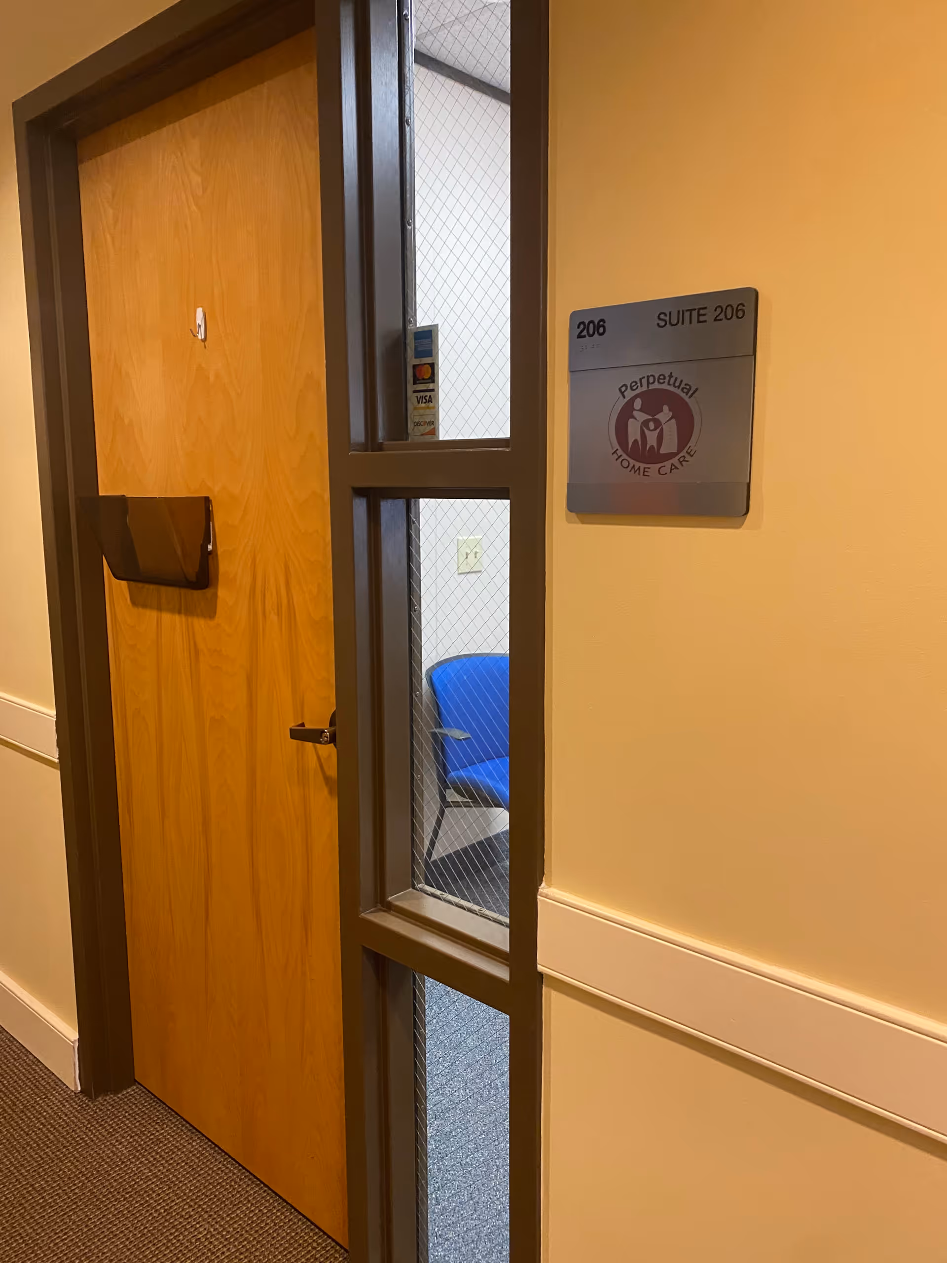 Hallway with a wooden door and glass window; blue chair visible beyond, Perpetual Home Care sign.