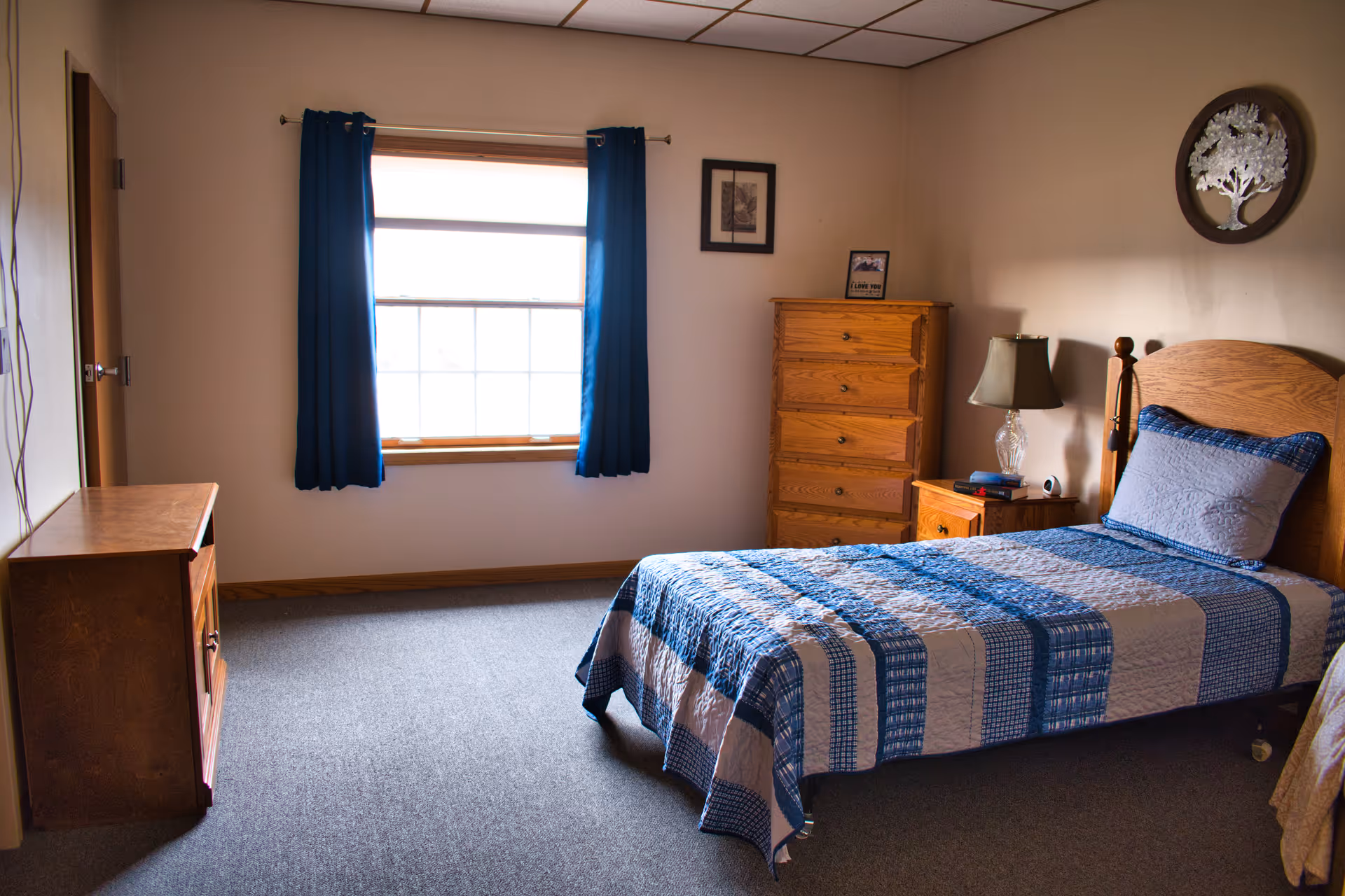 A tidy bedroom with a single wooden bed, blue quilt, dresser, and window with blue curtains.