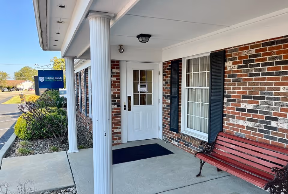Front entrance of a brick senior facility with a white door, shutters, and a red bench.