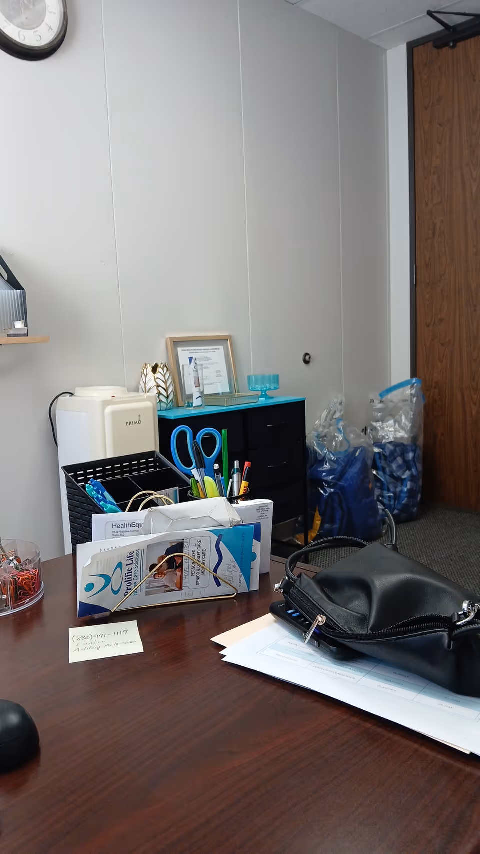 Organized office desk with files, stationery, and a black purse in a small facility office.