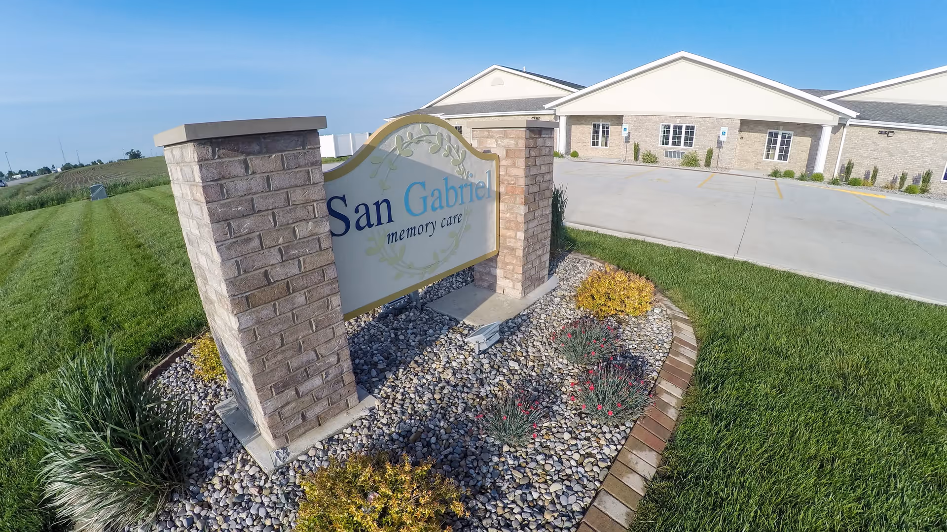 A brick monument sign that reads 'San Gabriel memory care' set in a rock and plant bed with the facility building and parking area behind it.