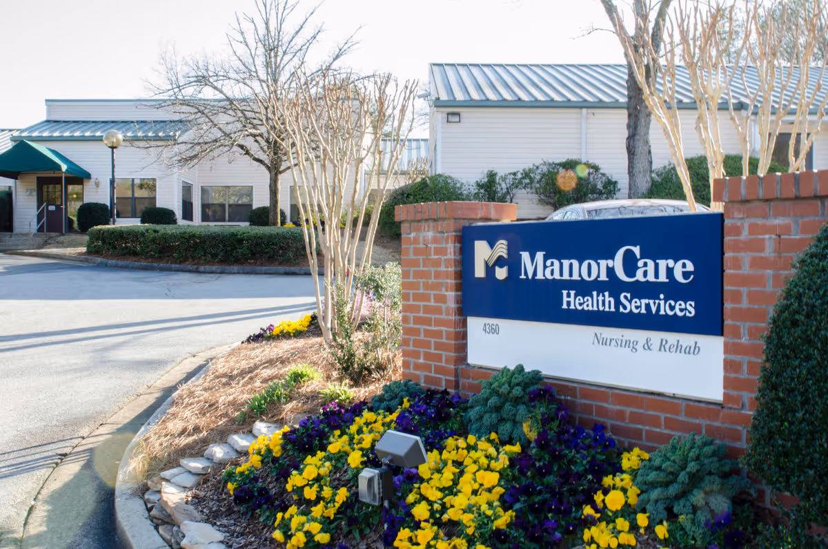 Exterior view of ManorCare Health Services building with a brick sign in front that reads 'ManorCare Health Services Nursing & Rehab'. The area around the sign is landscaped with yellow and purple flowers, small shrubs, and trees without leaves. The building has a light-colored exterior with a metal roof and a green awning over the entrance.