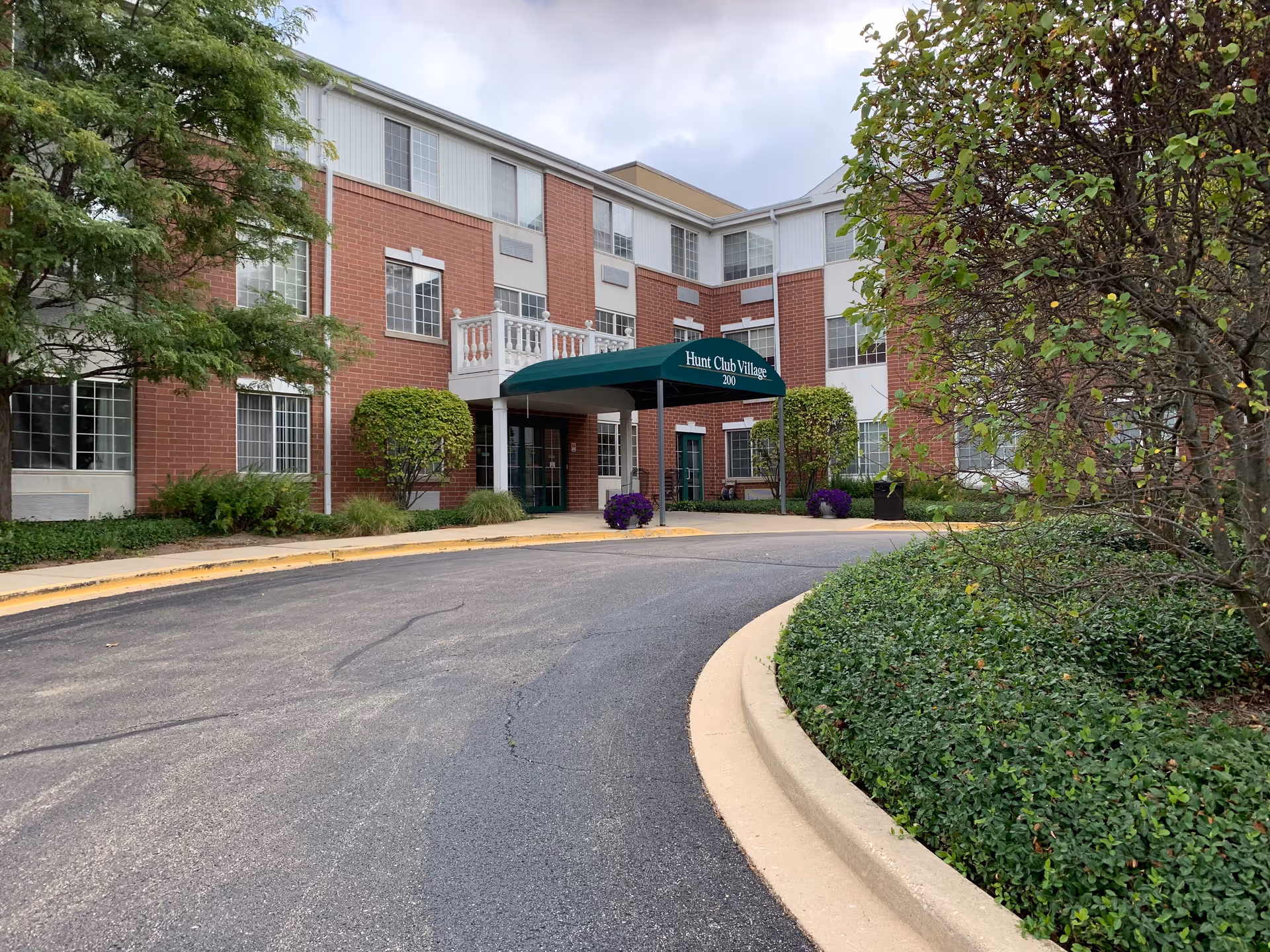 Exterior view of Hunt Club Village Senior Apartments showing a three-story brick and white panel building with a green awning over the entrance. There is a curved driveway leading to the entrance, surrounded by greenery including bushes and trees under a cloudy sky.