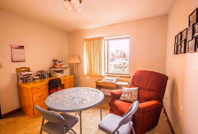 A small, cozy room with a round table covered by a lace tablecloth, surrounded by three chairs including a red cushioned armchair. There is a wooden desk with a lamp, books, and a printer against one wall, and a window with beige curtains letting in natural light. Several framed photos are hung on the wall to the right.