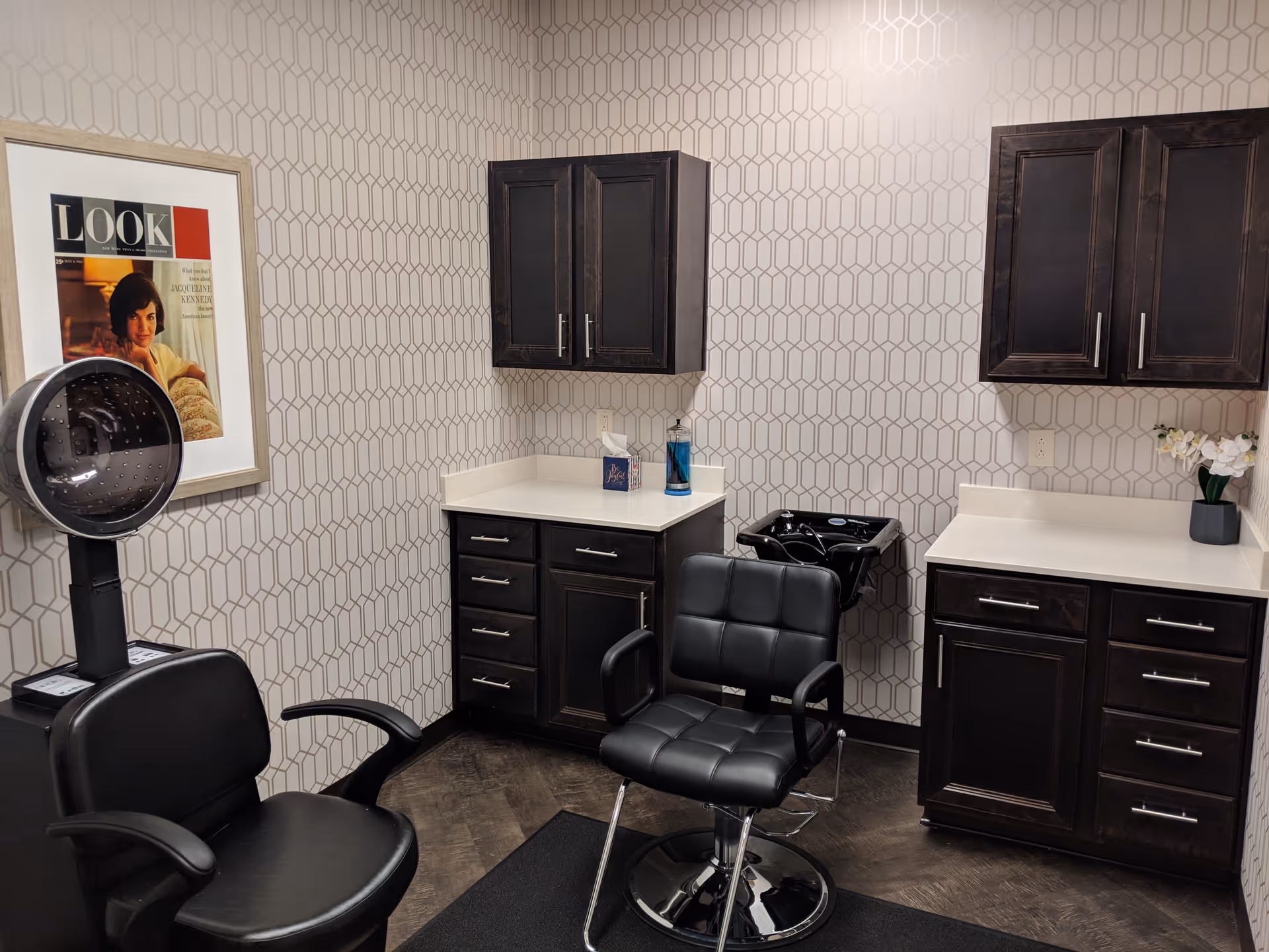 Hair salon area with dark wood cabinets, black styling chairs, a hooded dryer, and geometric wallpaper.