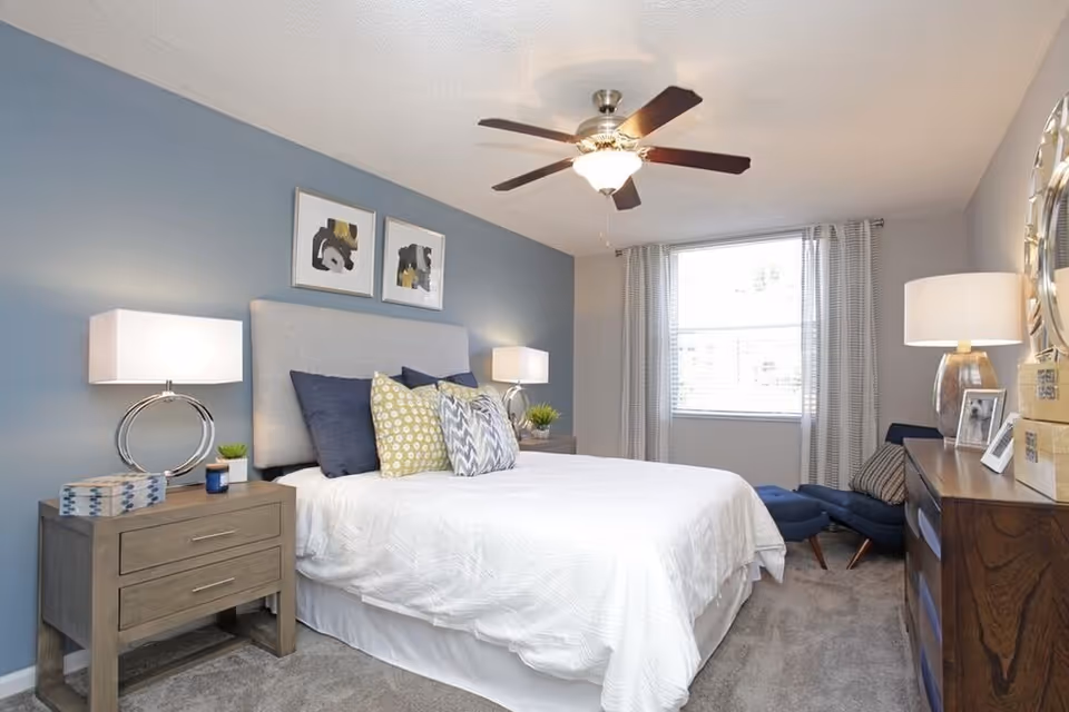 Bright bedroom with a bed dressed in white, nightstands and lamps, decorative pillows, a ceiling fan, and a window with curtains.