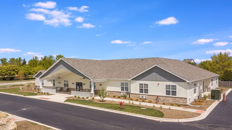 Single-story light-colored senior living building with a covered front entrance, landscaping, and a paved driveway under a blue sky.