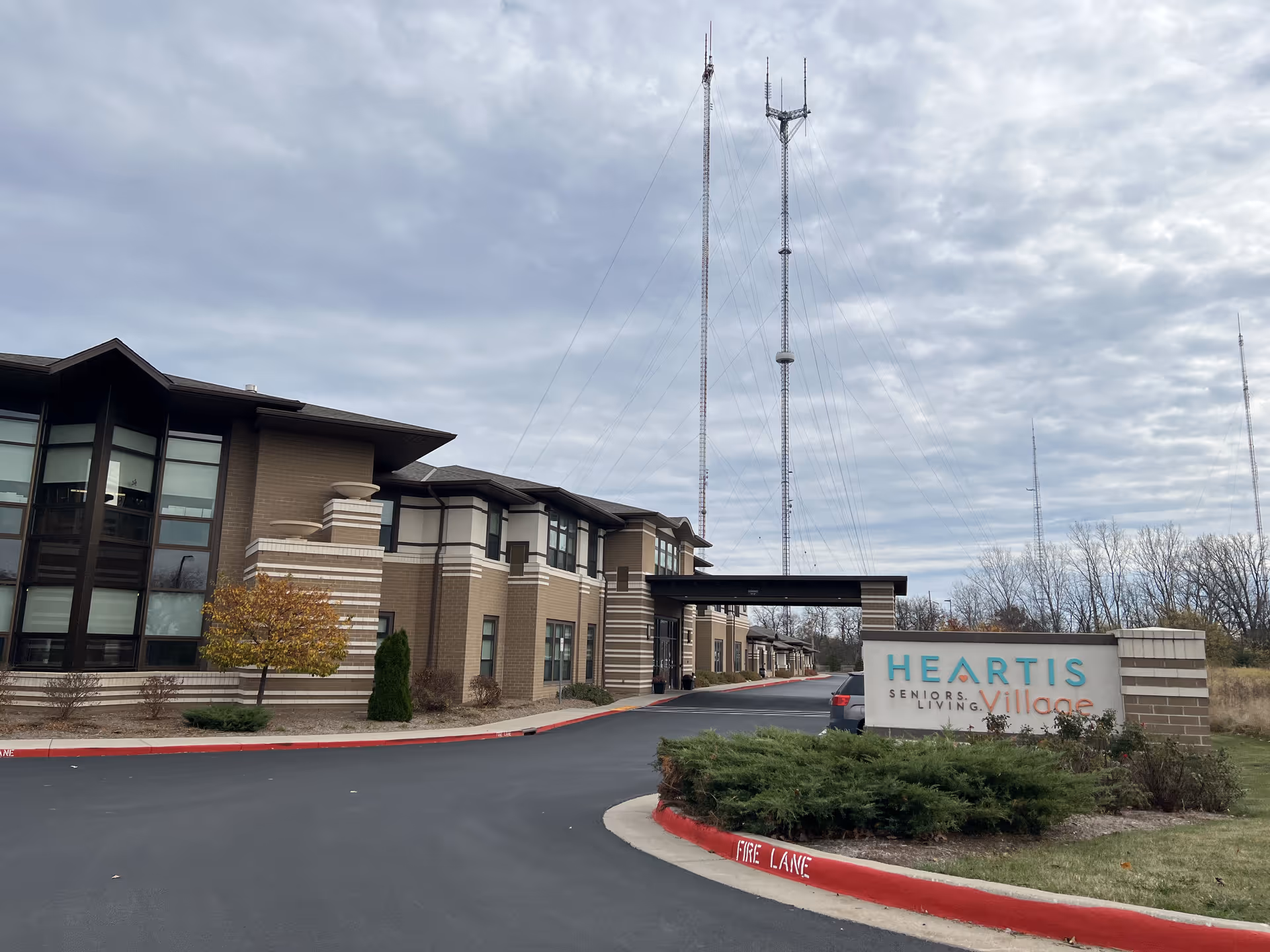 Exterior view of a senior living facility building with a driveway and a sign that reads 'Heartis Seniors Living Village'. The building is two stories with large windows and brick accents. There are tall communication towers in the background and some trees and shrubs around the property under a cloudy sky.