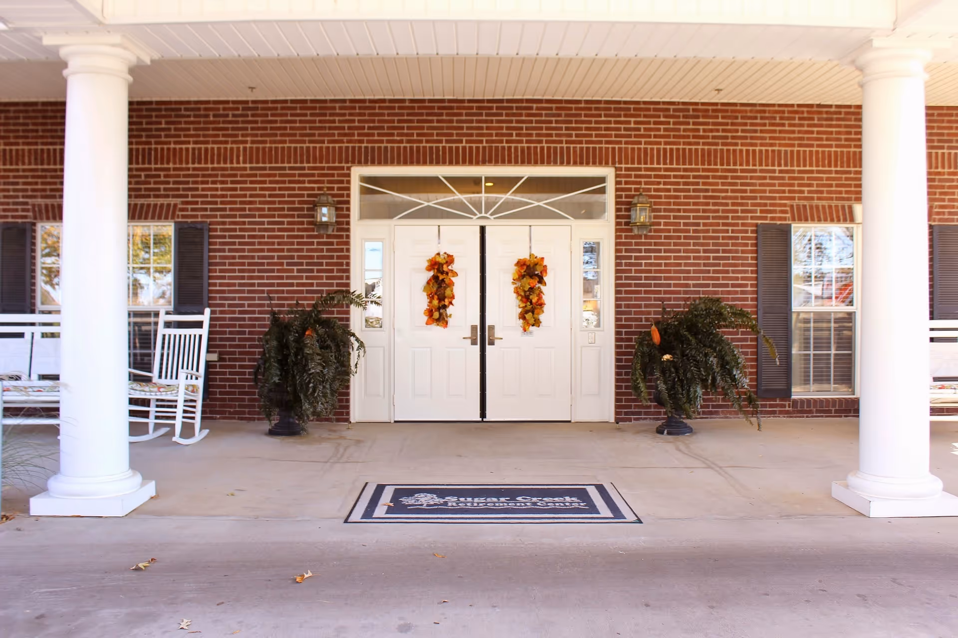 Entrance of a senior living facility with white double doors decorated with fall-themed wreaths, flanked by two large potted plants. The building has a red brick exterior with white columns and rocking chairs on the porch. A welcome mat with the facility name 'Sugar Creek Senior Living' is placed in front of the doors.