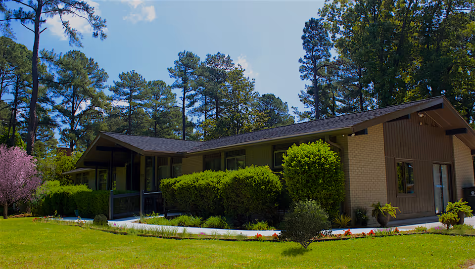 Single-story building with a sloped roof surrounded by green bushes and trees under a blue sky with some clouds. The building has large windows and a small porch area. The lawn in front is well-maintained with some flowers and small plants.