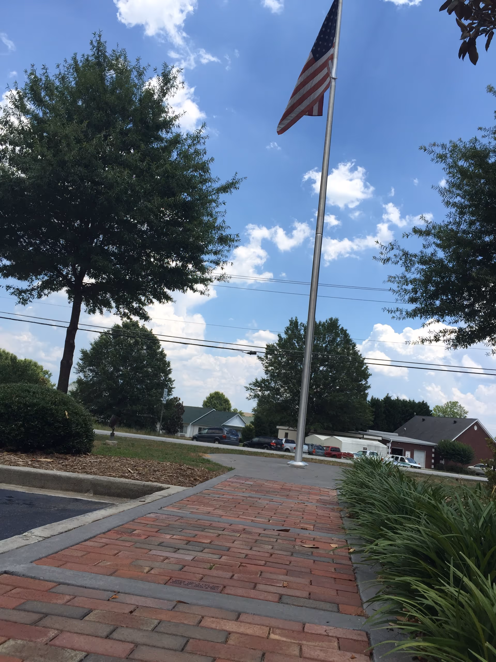 American flag on a tall pole beside a brick walkway, trees, and parked cars.