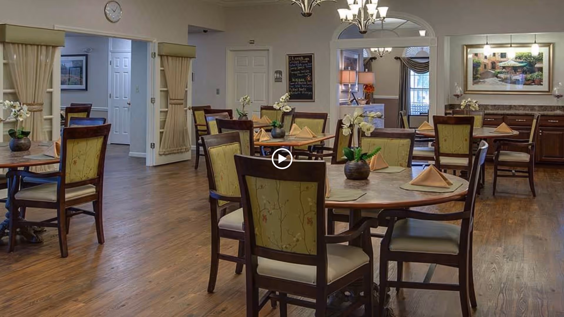 A dining room in a senior living facility with round tables set with folded napkins and small flower arrangements. The room has wooden floors, upholstered chairs, and soft lighting. There is a chalkboard menu on the wall and a framed picture above a sideboard in the background.