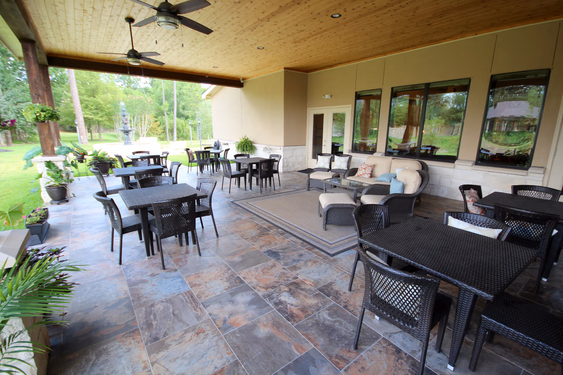 Covered outdoor patio with black wicker tables, chairs, and a beige sofa set overlooking a lawn.