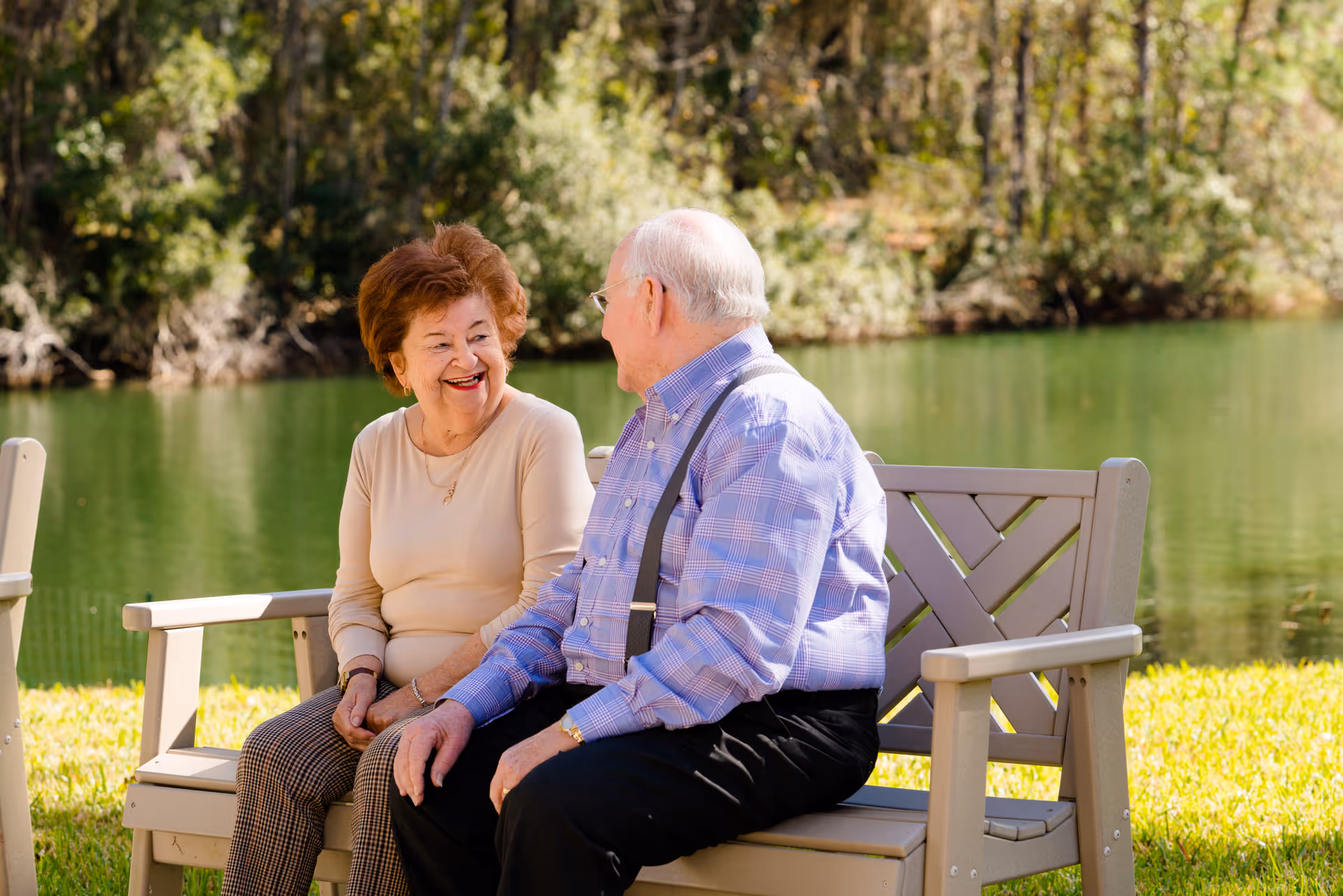 Two elderly residents sit on a bench by a lake, smiling and chatting outdoors.