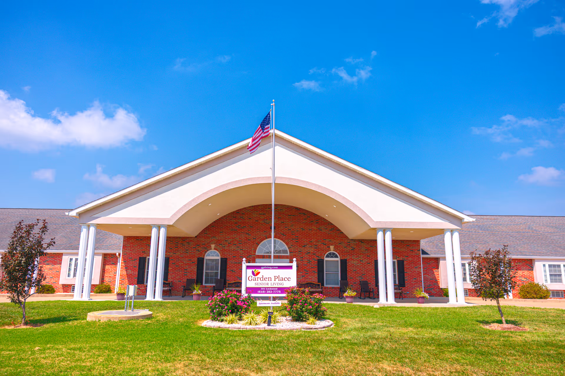 Brick exterior of a senior living facility with arched entry and flag on a sunny day.