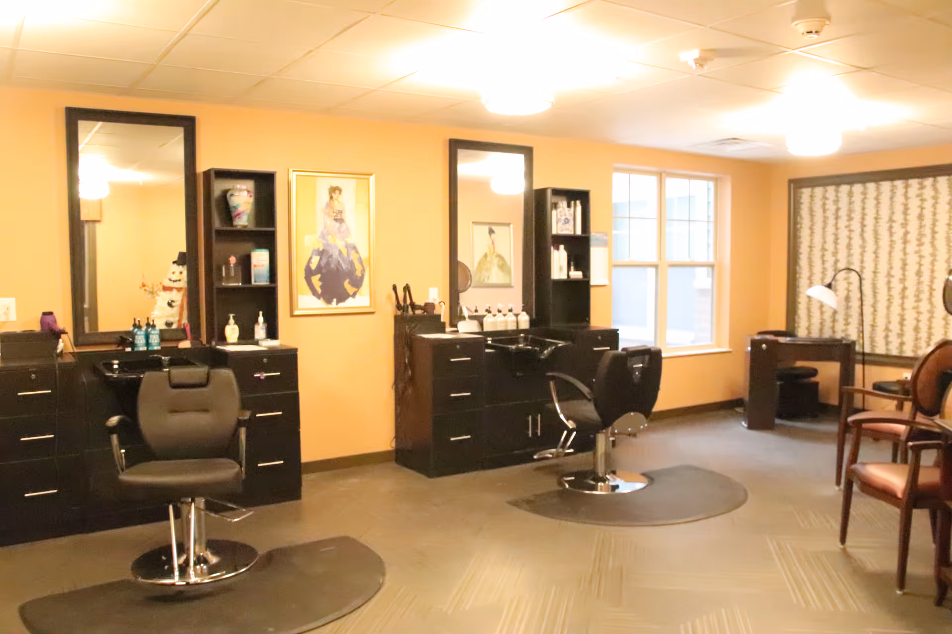 Interior view of a salon area in a senior living facility with two black salon chairs in front of mirrors and cabinets. The room has warm yellow walls, framed artwork, a window letting in natural light, and additional seating with chairs and a small desk in the background.