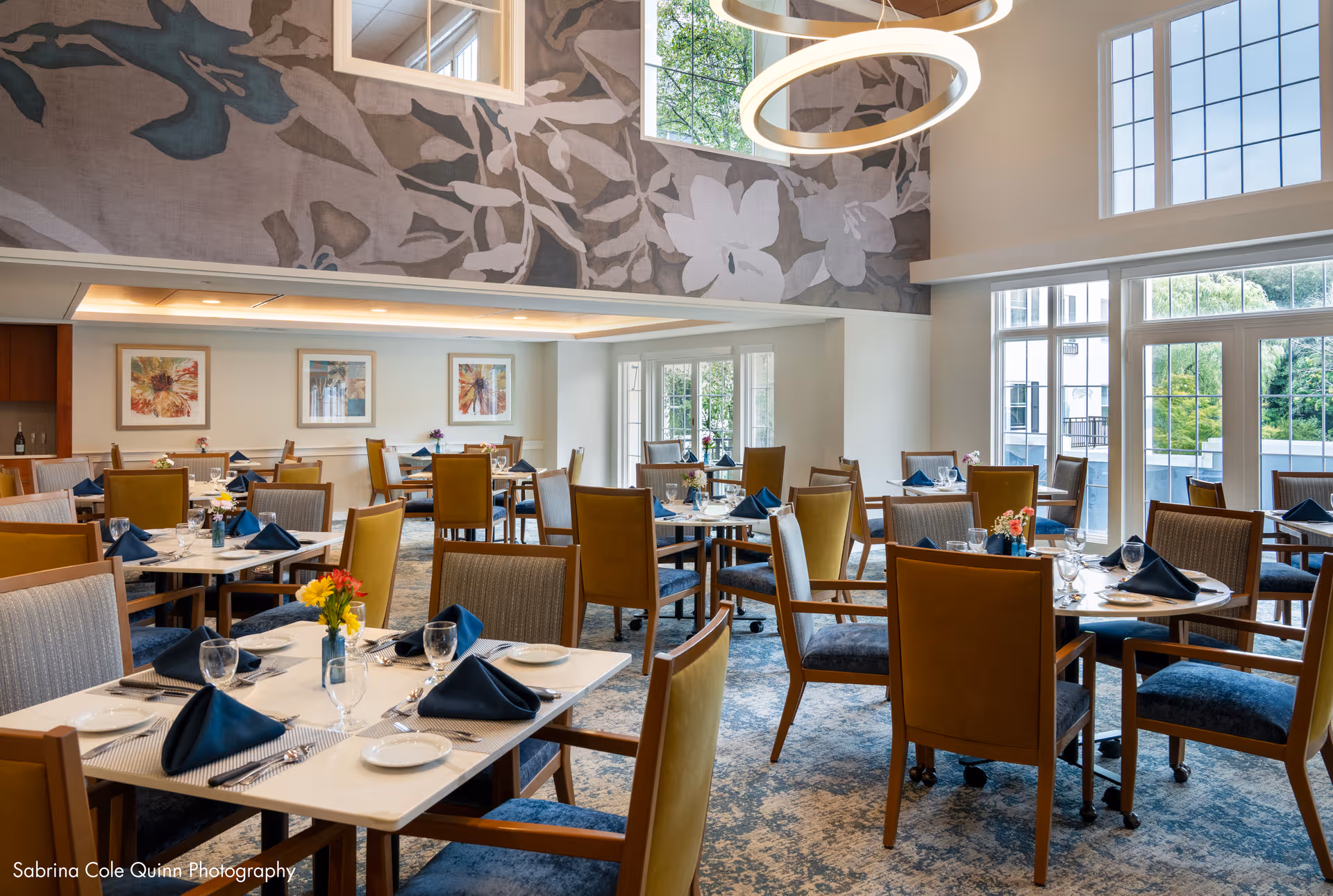 Bright senior dining room with many tables set for meals, flowers, and large windows.
