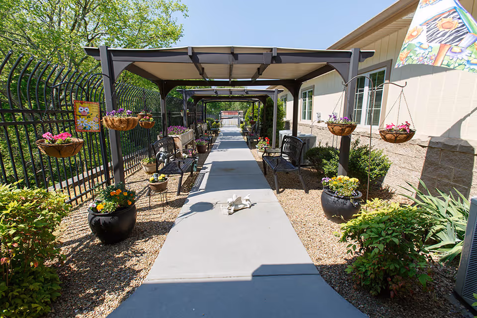 Covered outdoor walkway alongside a building with benches, hanging flower baskets and potted plants.