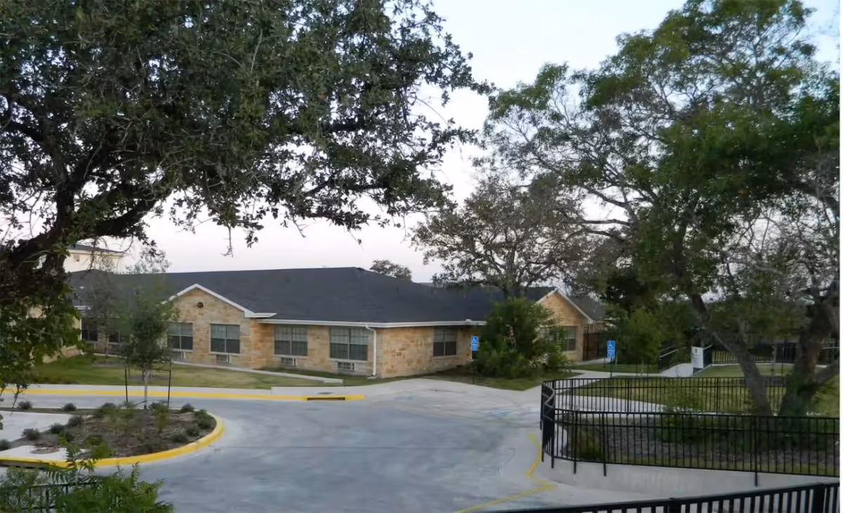 Single-story stone nursing facility building seen through trees with a curved driveway and parking area in front.