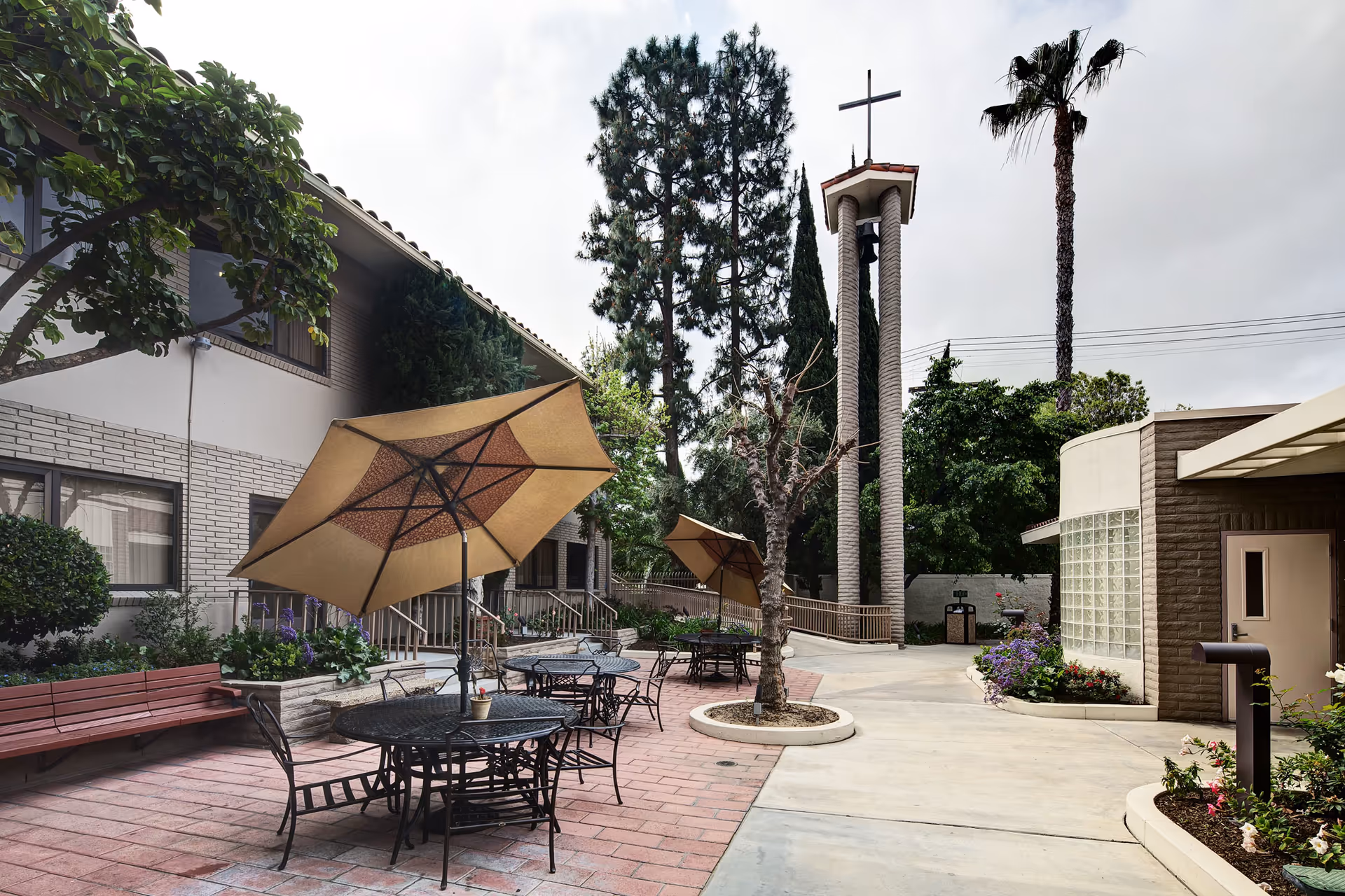A courtyard patio with round metal tables and umbrellas, benches, trees and a tall bell tower topped with a cross beside the building.