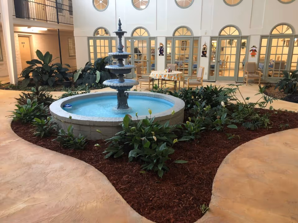 Indoor atrium with a tiered fountain surrounded by plants and seating in front of arched windows and doors.