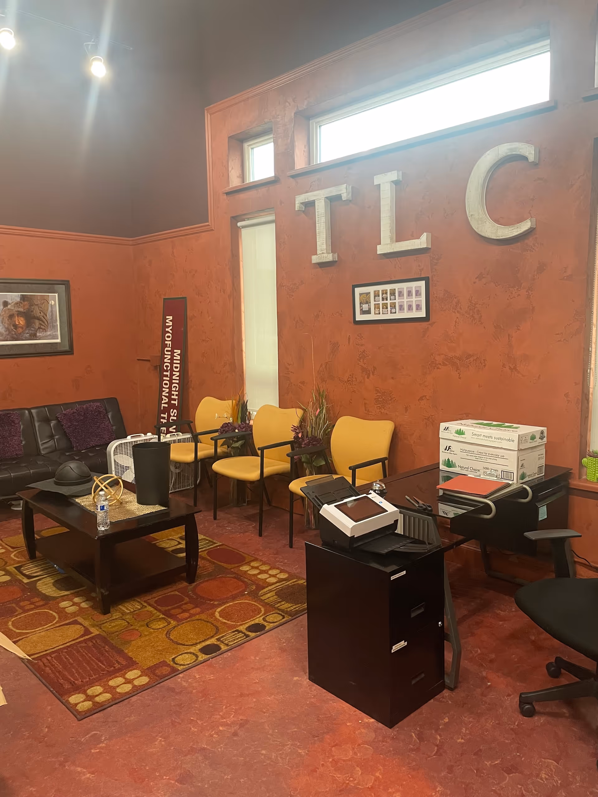 Interior lounge area with orange walls, yellow chairs, sofa, coffee table, and TLC signage.