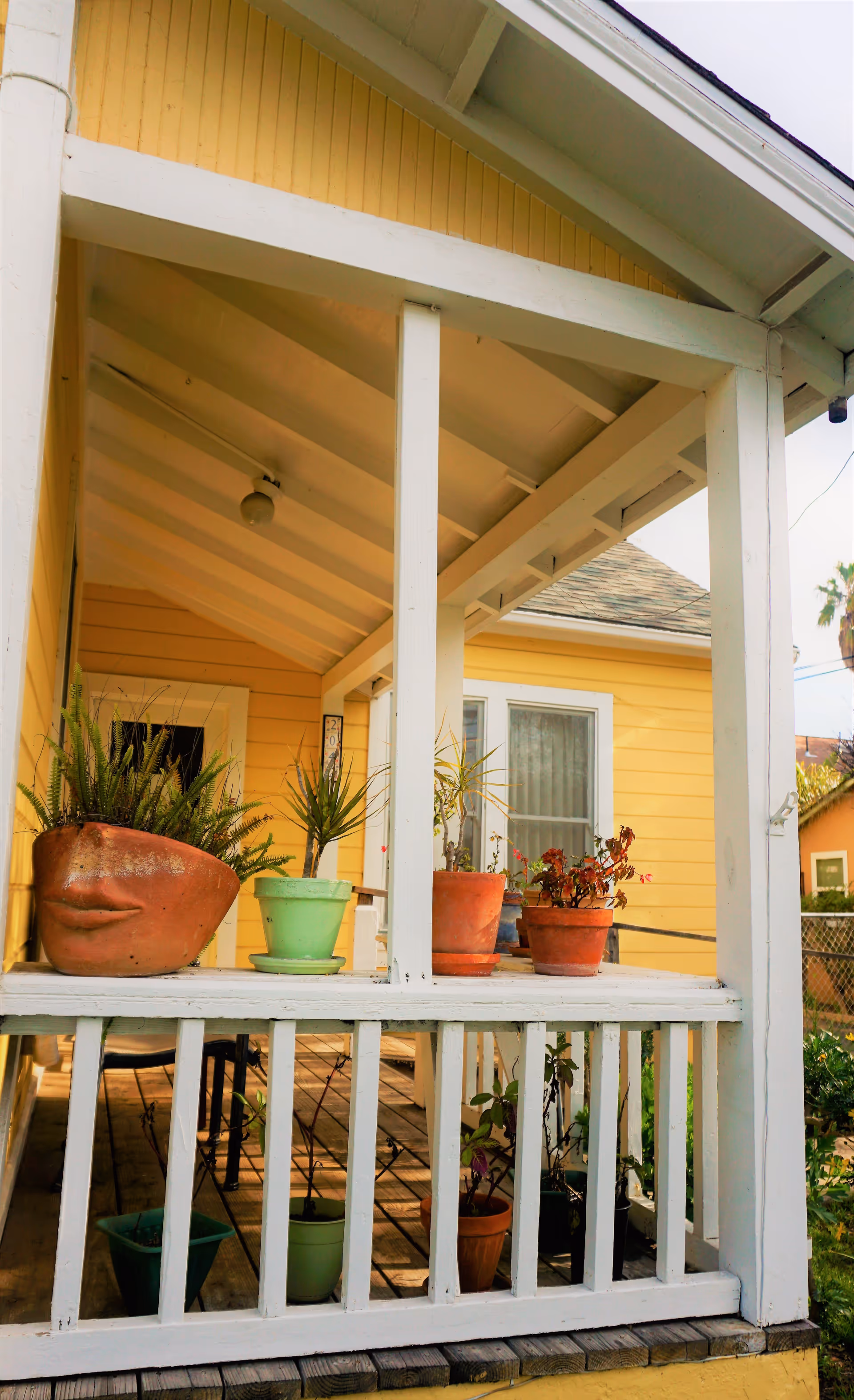 A yellow house porch with white railings and beams, featuring several potted plants on the railing and floor. The porch has a wooden floor and a slanted roof with a light fixture.