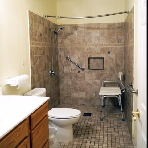 Accessible bathroom with tiled walk-in shower, shower chair, grab bars, toilet, and sink.