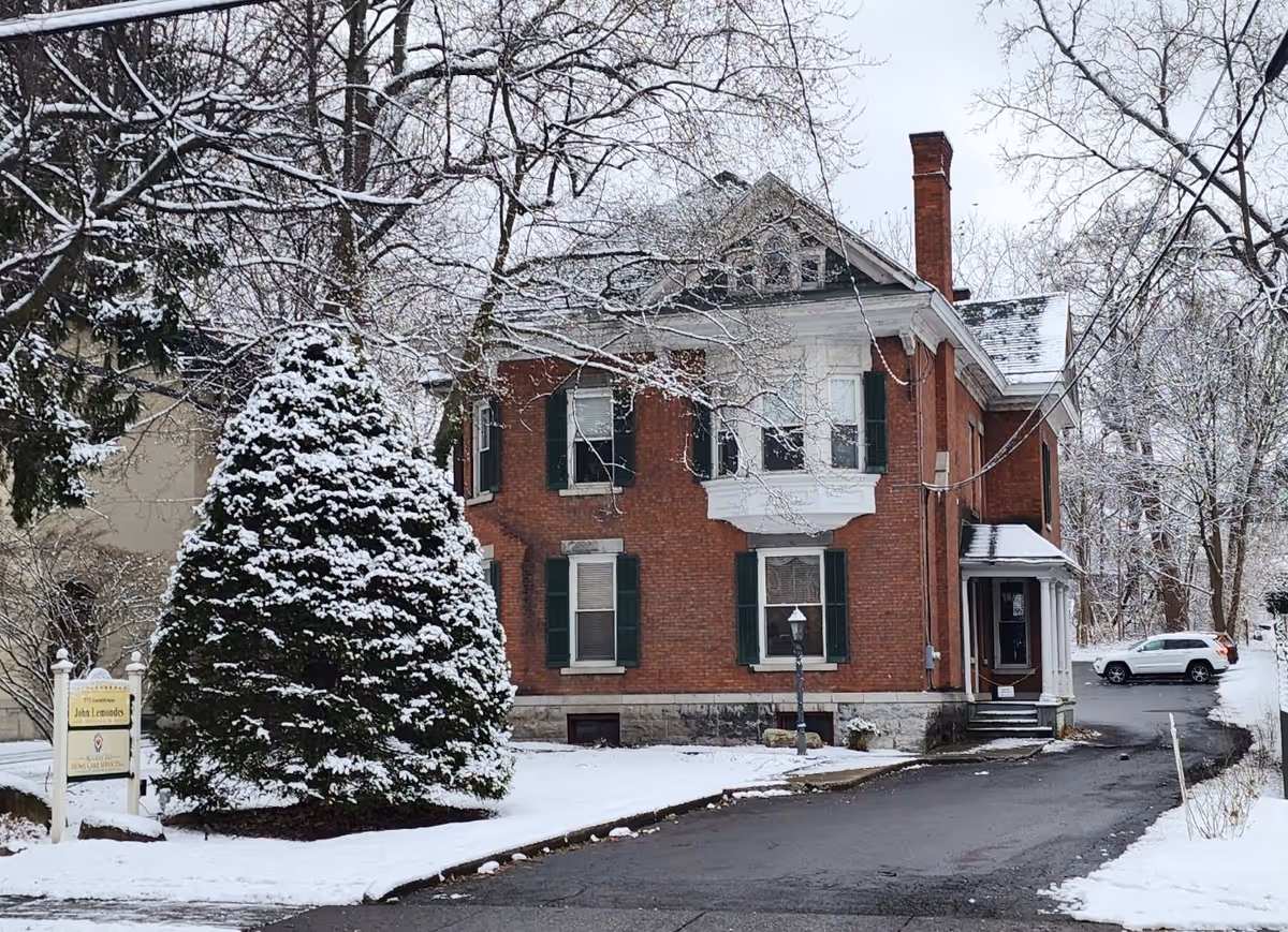 Snow-covered brick two-story building with driveway, trees, and a small front sign.
