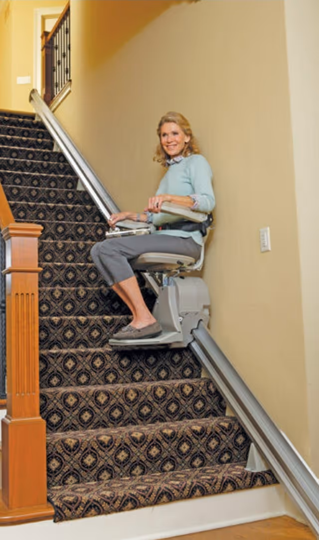A woman sits on an indoor stairlift along a carpeted staircase.