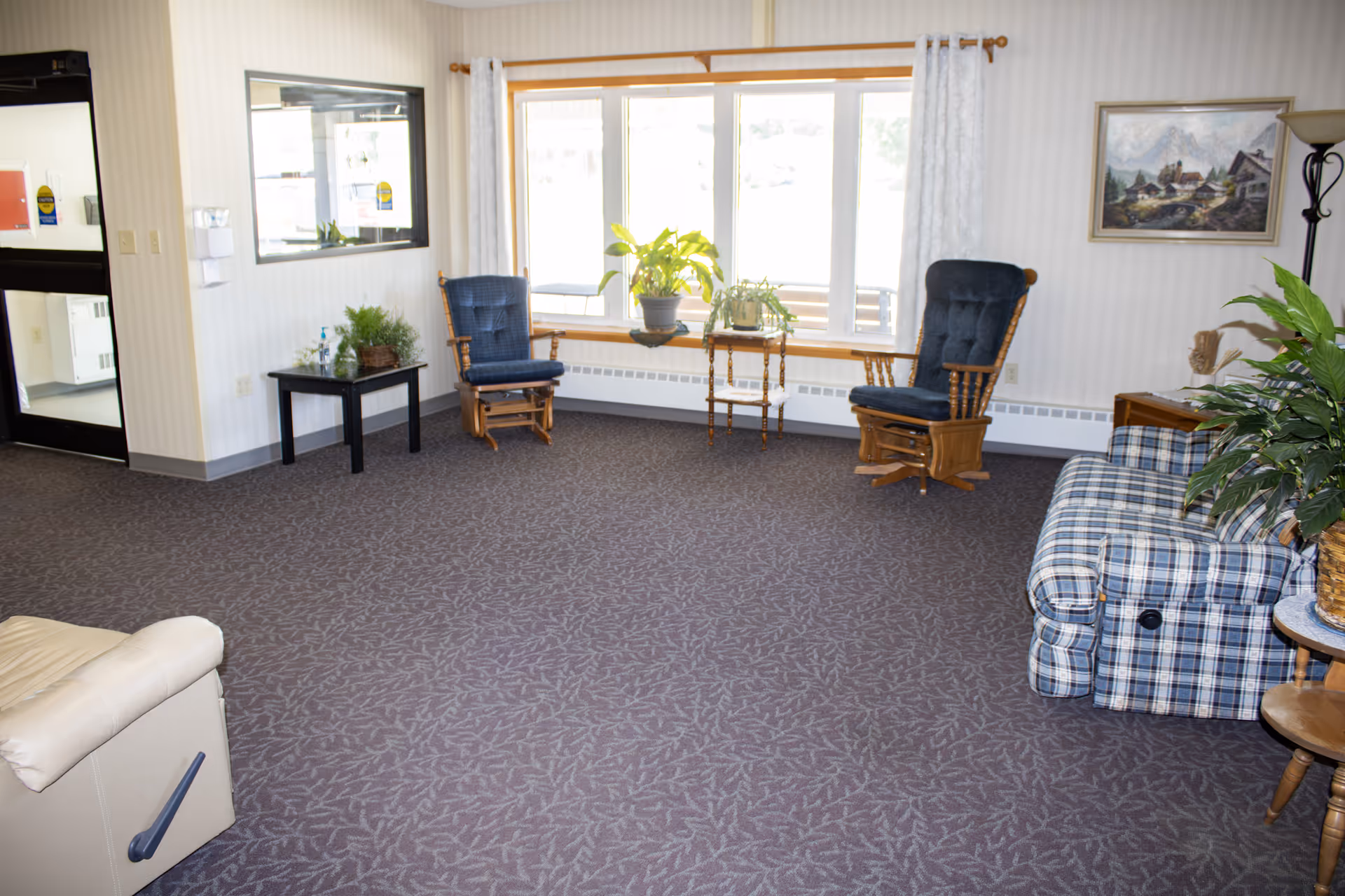 Cozy senior living lounge with blue chairs, plaid sofa, plants, and large front windows.