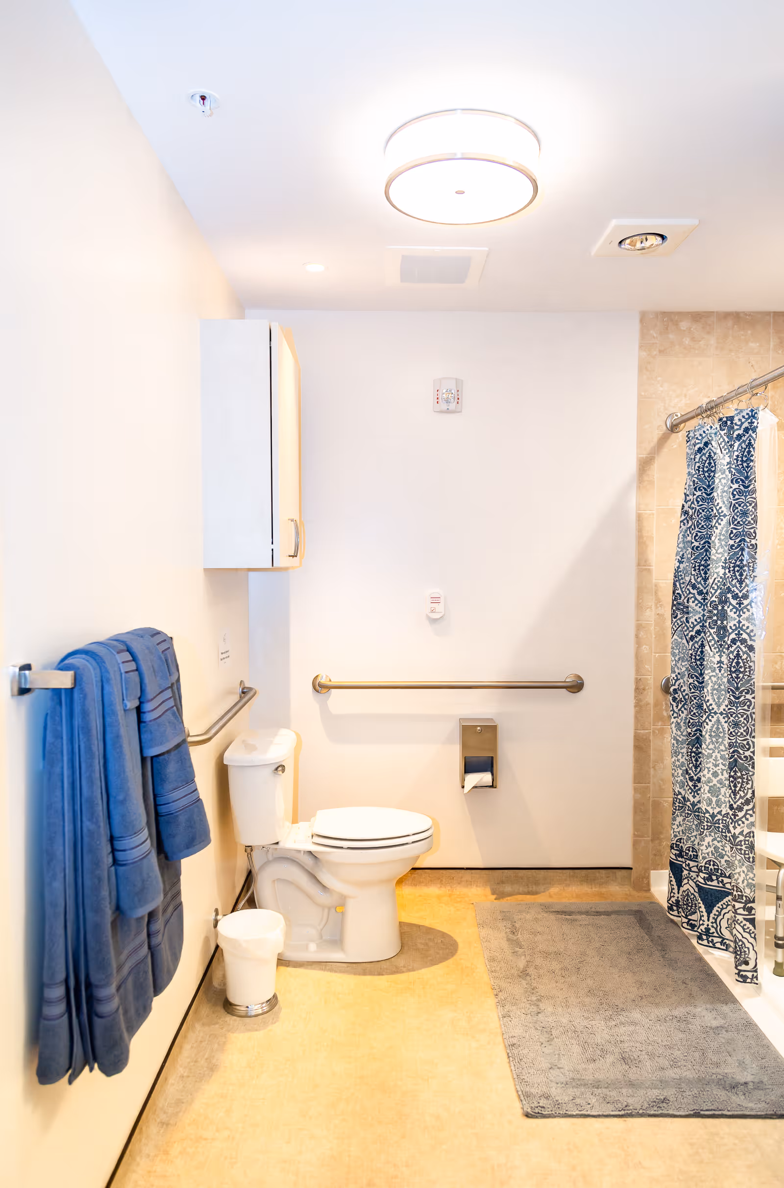 Accessible bathroom with a toilet, wall grab bars, blue towels on a rack, and a shower with a patterned curtain.