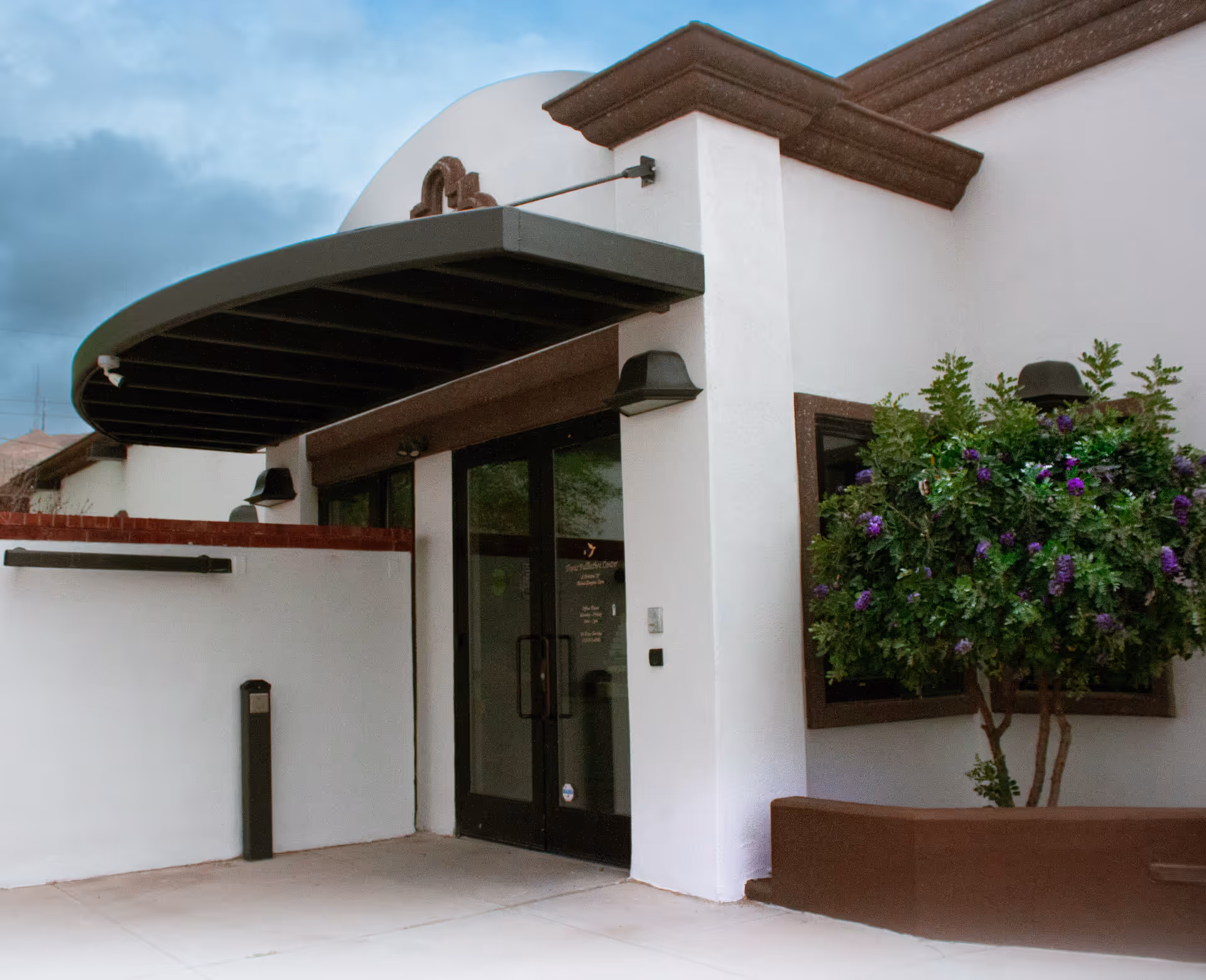 White stucco building entrance with curved dark awning and purple flowering tree nearby.