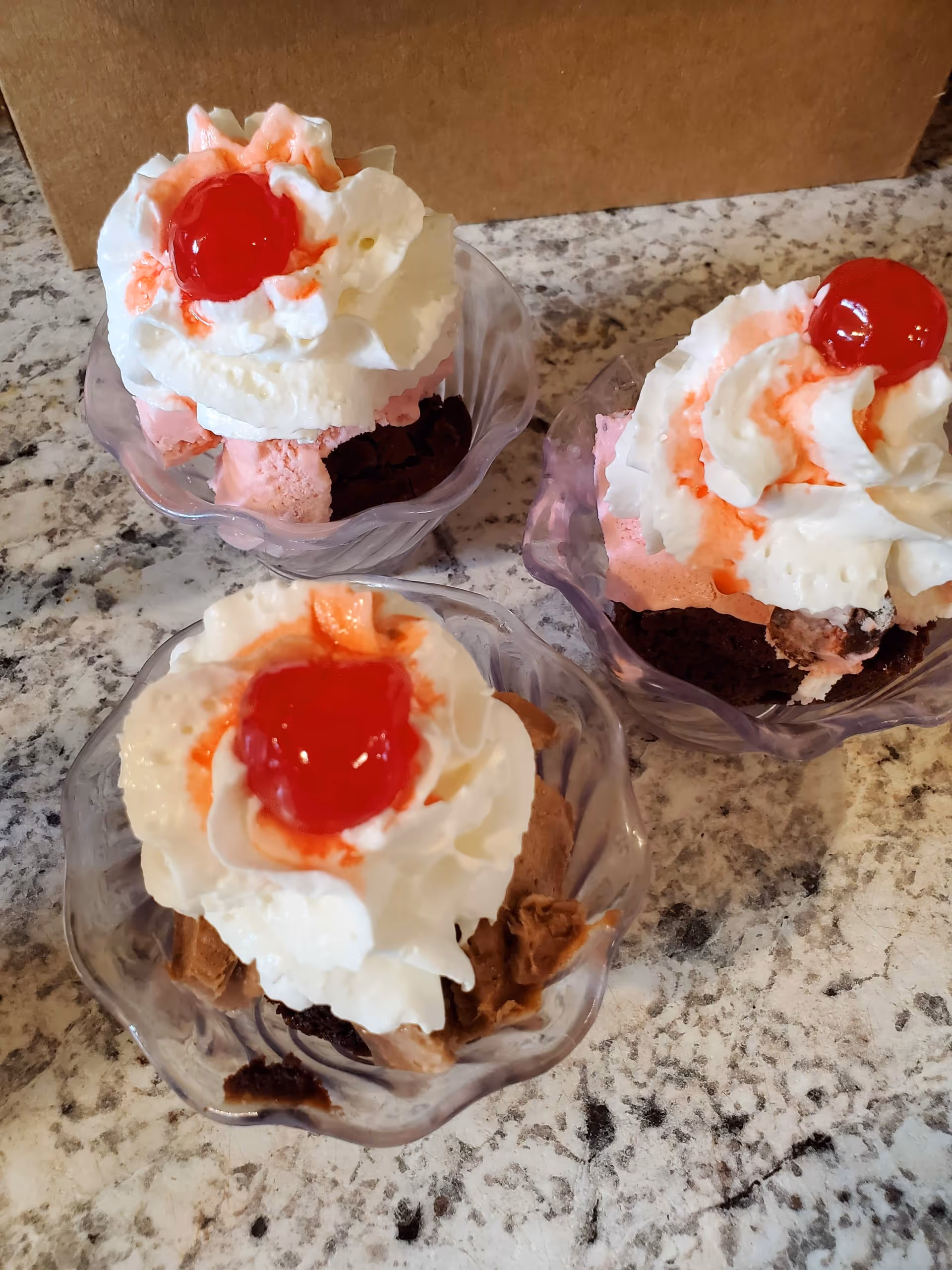 Three glass dessert cups with whipped cream and cherries sit on a granite counter.