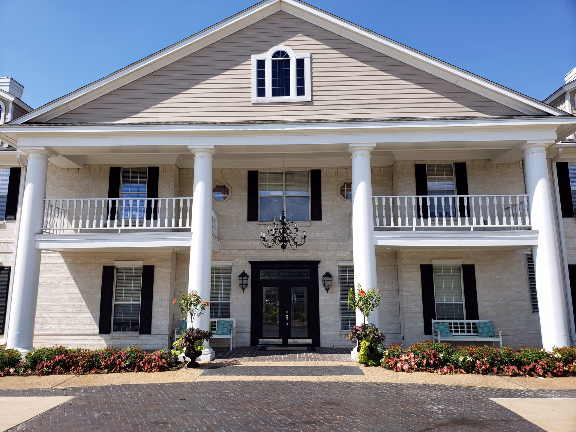 Front exterior view of a large assisted living facility building with beige brick walls, white columns, black shutters on windows, and a black double door entrance. There are two balconies above the entrance and decorative plants and benches on either side of the door under a clear blue sky.