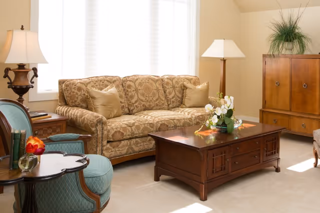 Well-lit living room with a patterned sofa, wooden coffee table with flowers, upholstered armchair, lamps, and a wooden cabinet.
