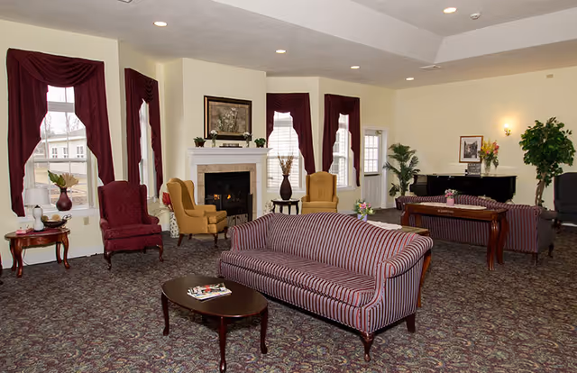 Communal living room with striped sofas and armchairs arranged around a fireplace, a piano, and burgundy window drapes.