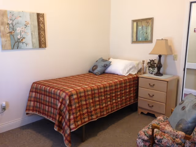 Single bedroom with plaid bedspread, nightstand, lamp, and floral chair.