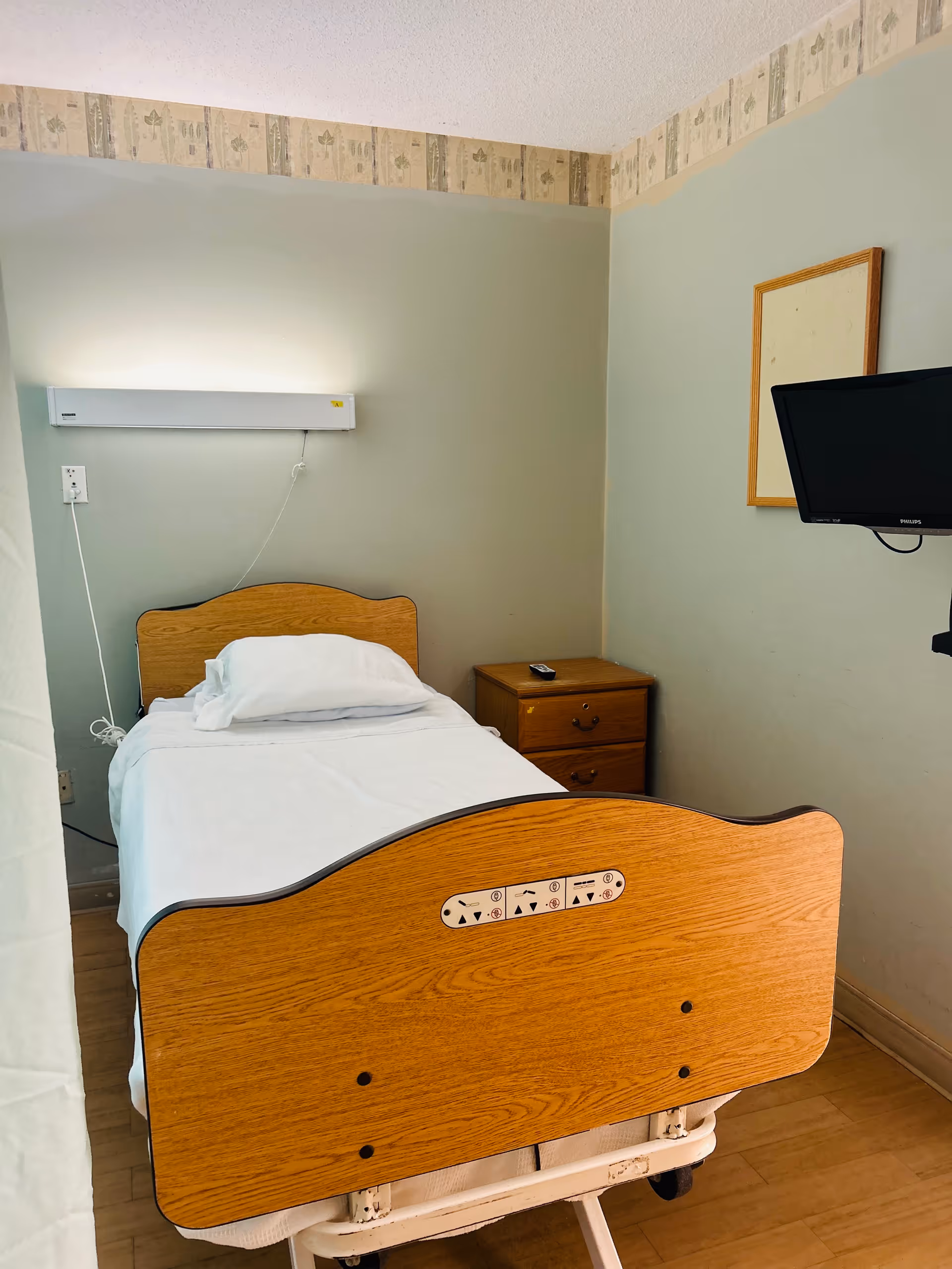 A small, simple room with a single hospital-style bed with wooden headboard and footboard, a white pillow and sheet, a wooden nightstand with a remote control on top, a wall-mounted flat screen TV, and a light fixture above the bed. The walls are painted light green with a decorative wallpaper border near the ceiling.